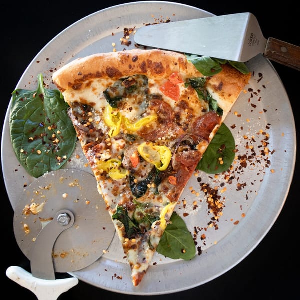 A slice of East Hill Pizza with spinach, peppers, and red pepper flakes on a metal pan.