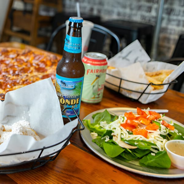 East Hill Pizza spread with pizza, salad, garlic knots, and two beers on a wooden table.
