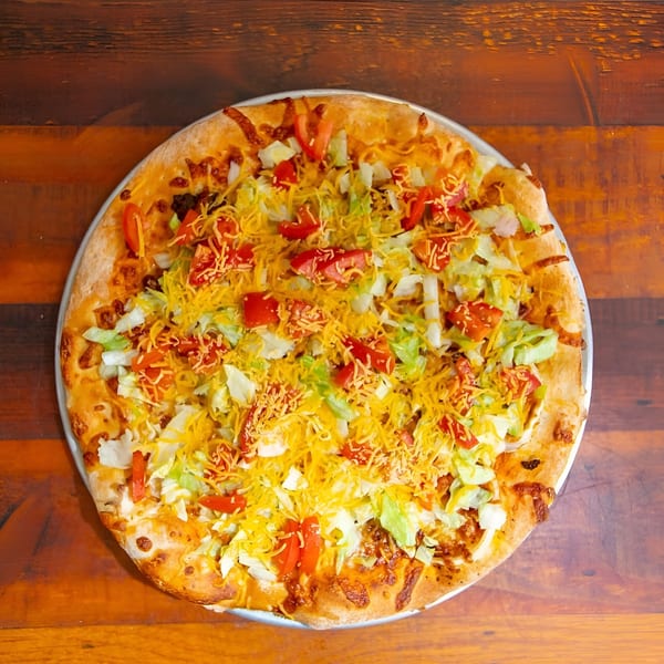 East Hill Pizza's taco pizza, topped with shredded cheese, lettuce, and diced tomatoes on a wooden table.