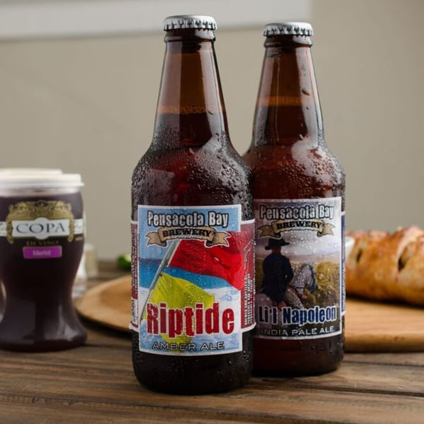 Two cold Pensacola Bay Brewery beer bottles, Riptide Amber Ale and Li'l Napoleon IPA, on a wooden table.