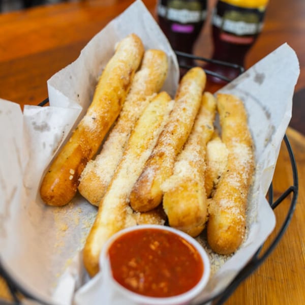 East Hill Pizza's cheesy breadsticks with marinara sauce in a basket, with drinks in the background.