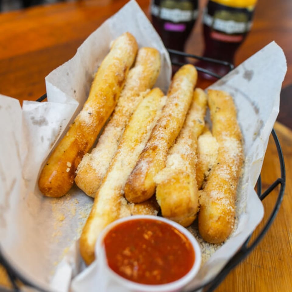 East Hill Pizza's cheesy breadsticks with marinara sauce in a basket, with drinks in the background.