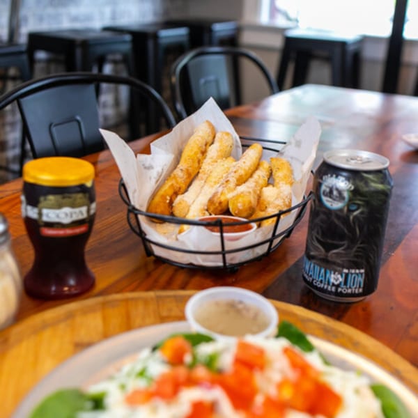 East Hill Pizza's cheesy breadsticks with marinara, a drink, and a pizza on a wooden table.